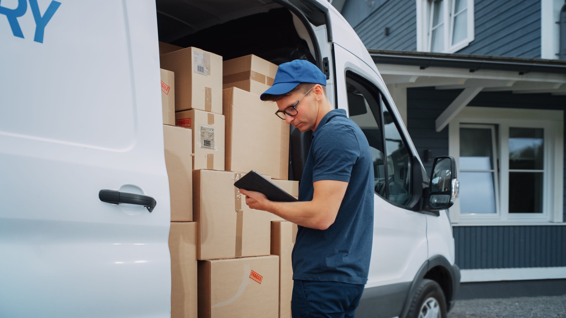 Courier Using Tablet Computer Next to Open Delivery Van Side Door with Cardboard Parcels. Mailman Delivering the Package to a Homeowner.